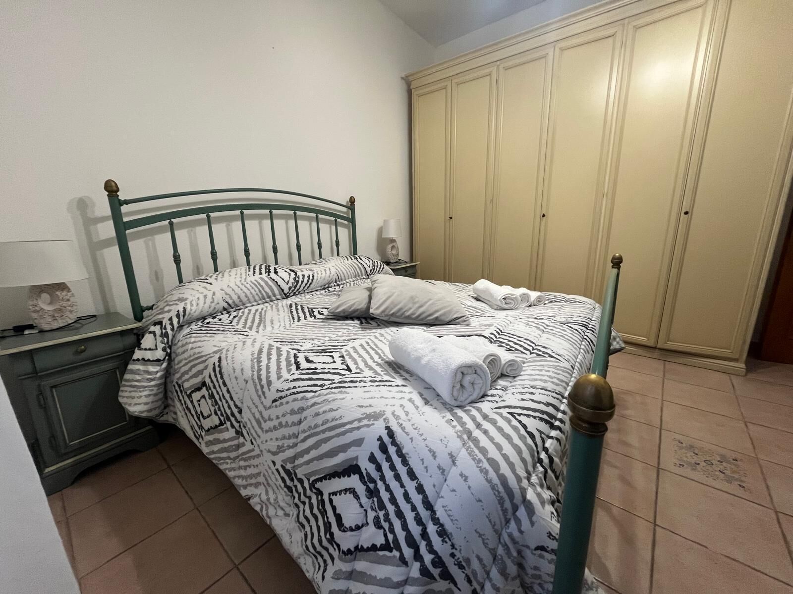 Uma cama com roupa de cama estampada, uma mesa de cabeceira com abajur e um guarda-roupa num quarto com chão em mosaico.