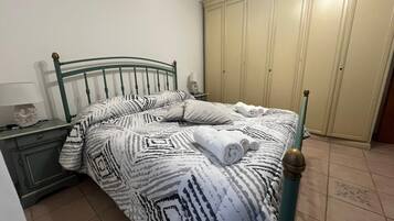 Uma cama com roupa de cama estampada, uma mesa de cabeceira com abajur e um guarda-roupa num quarto com chão em mosaico.