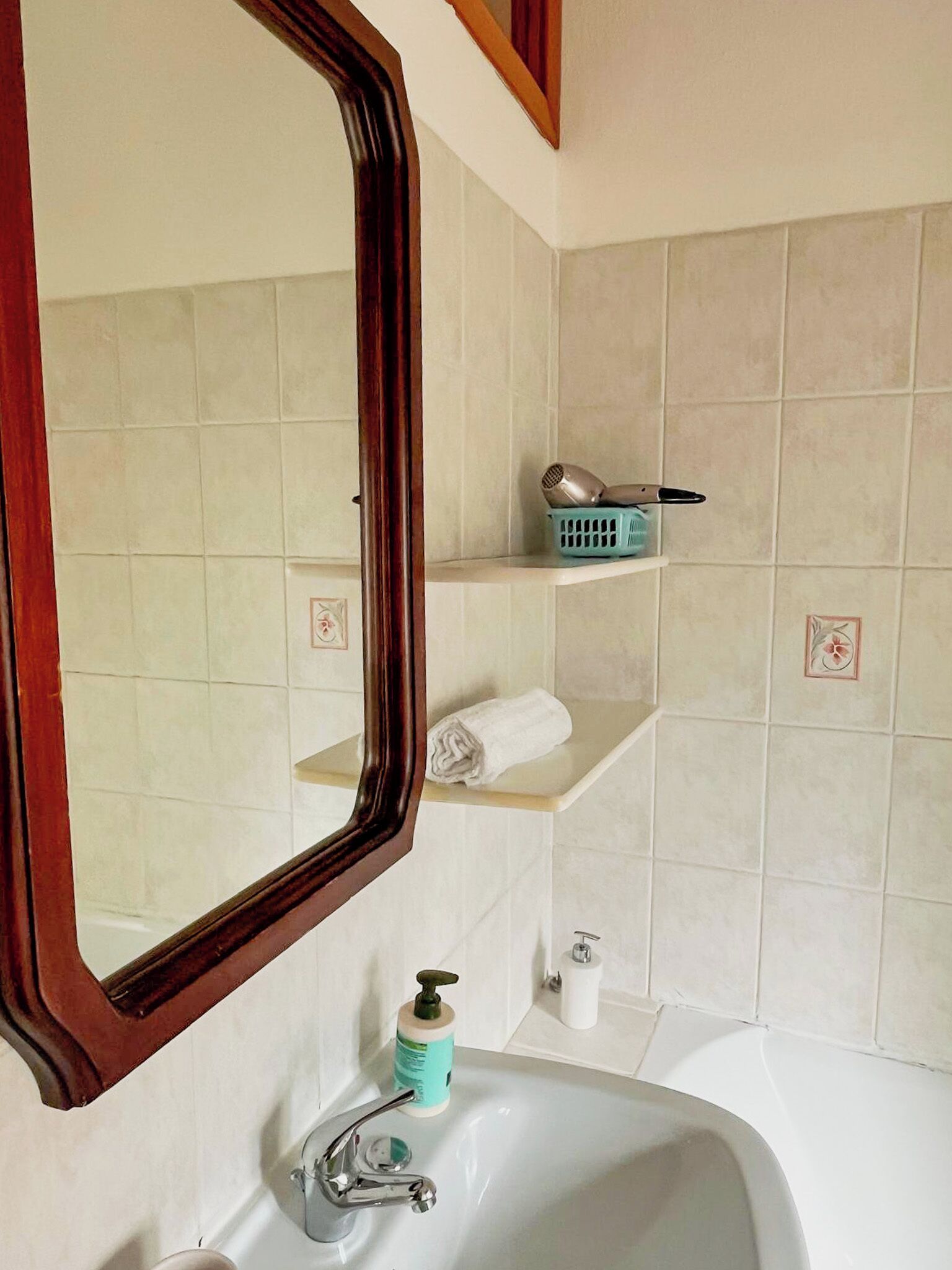 Ein Badezimmer mit einem weißen Waschbecken, einem Spiegel mit Holzrahmen, einem Handtuch, einem Fön und einer Seifenspender.