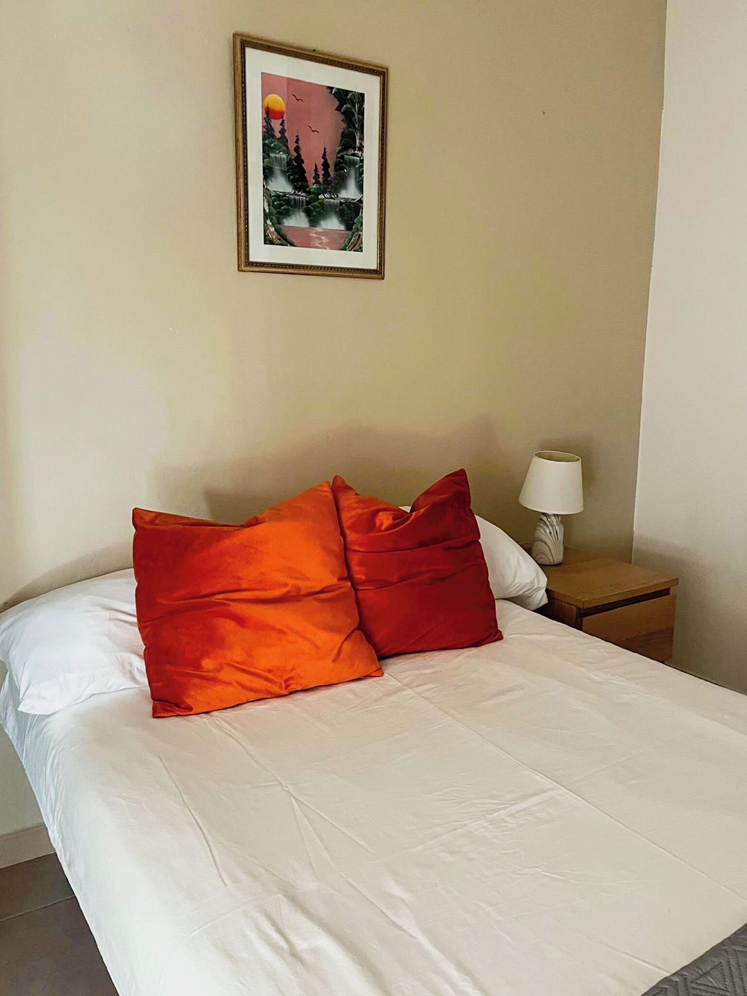 Una cama bien hecha con sábanas blancas y dos almohadas naranja brillante, un cuadro en la pared y una mesita de noche con una lámpara.