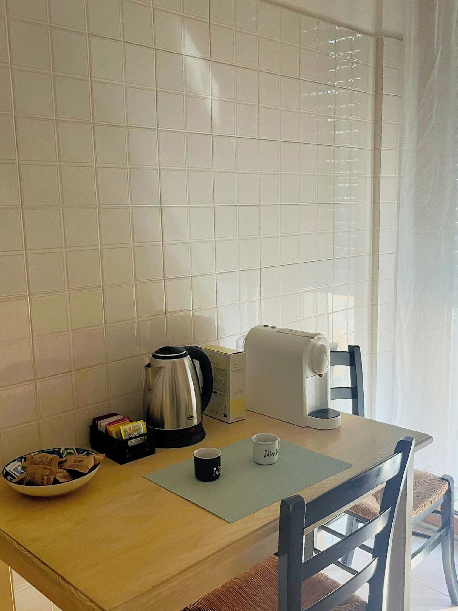 Et lille køkken med hvide fliser, en elkedel, en brødrister, en skål og en kop på et bord.