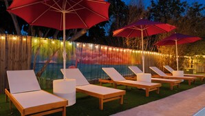 A poolside area with wooden lounge chairs and red umbrellas, surrounded by a wooden fence and string lights.