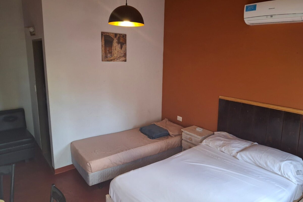 Hotelkamer met een eenpersoonsbed, een kleiner bed, een bureau met stoel, een hanglamp en een airconditioner aan de muur.