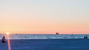 Escena de playa con el sol poniéndose sobre el agua, barcos y gente en la orilla.