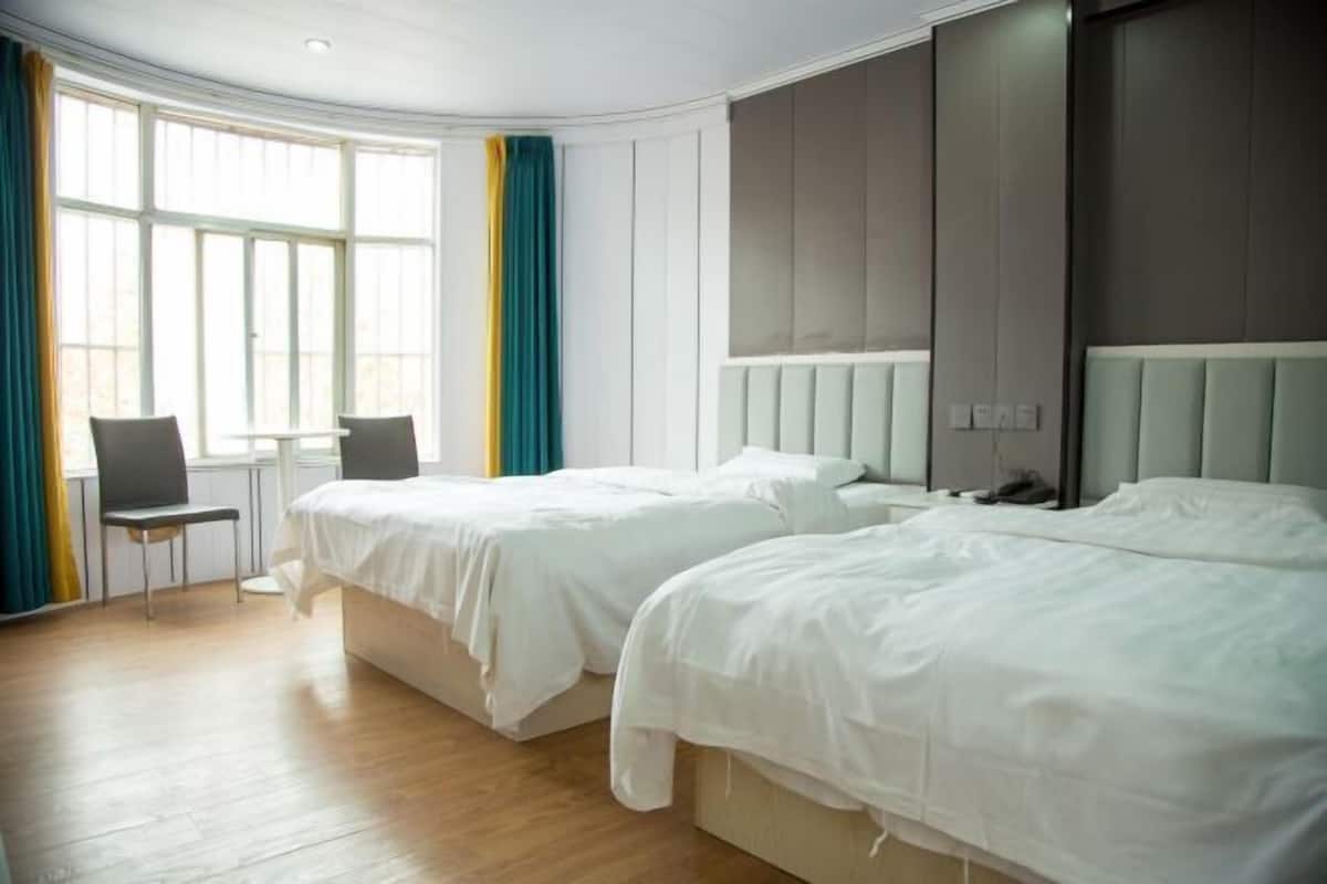 A hotel room with two beds, a chair, a window with curtains, and a light-colored wooden floor.