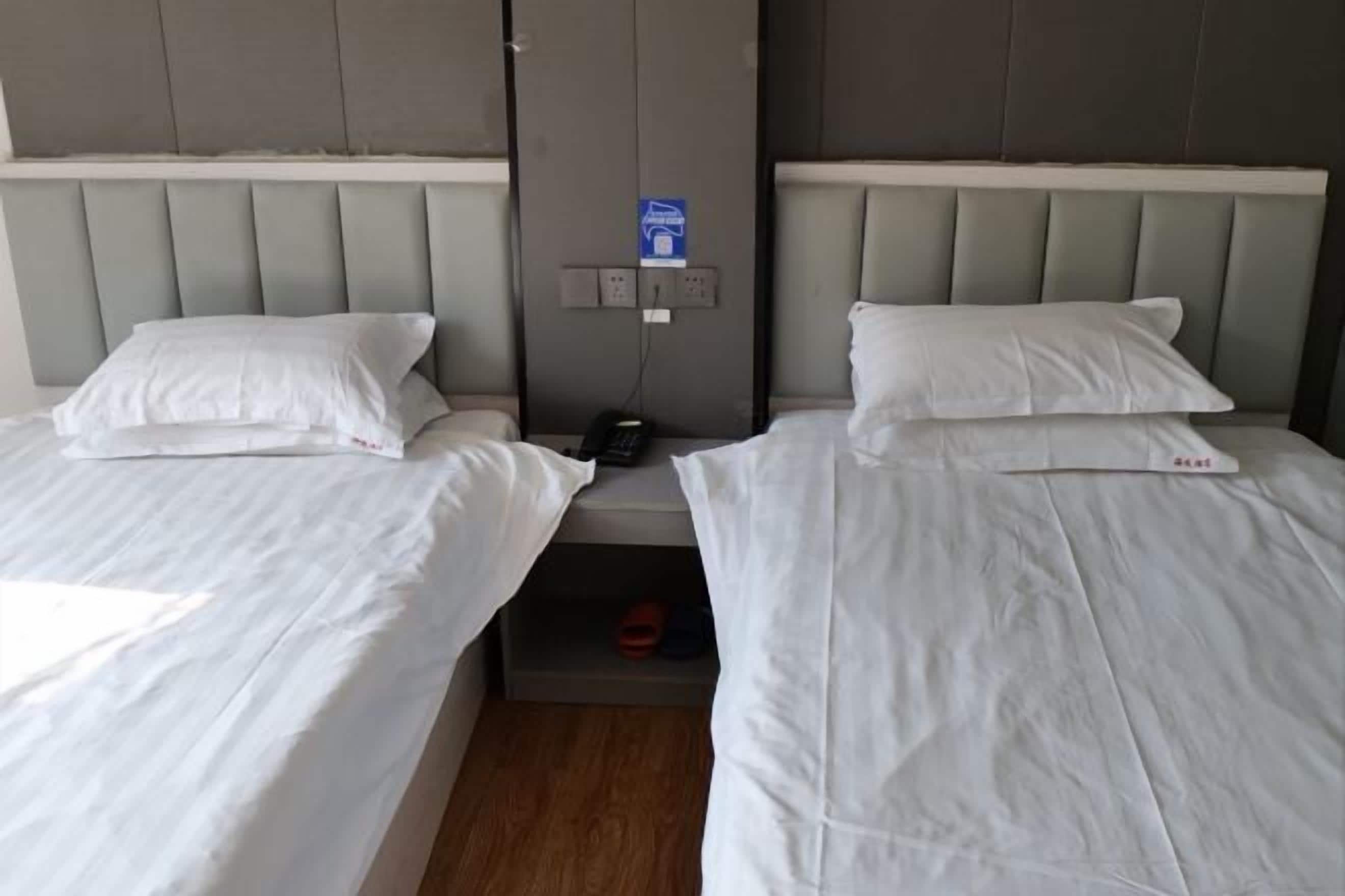 A hotel room with two single beds, a nightstand, a phone, and a small shelf with a red object.