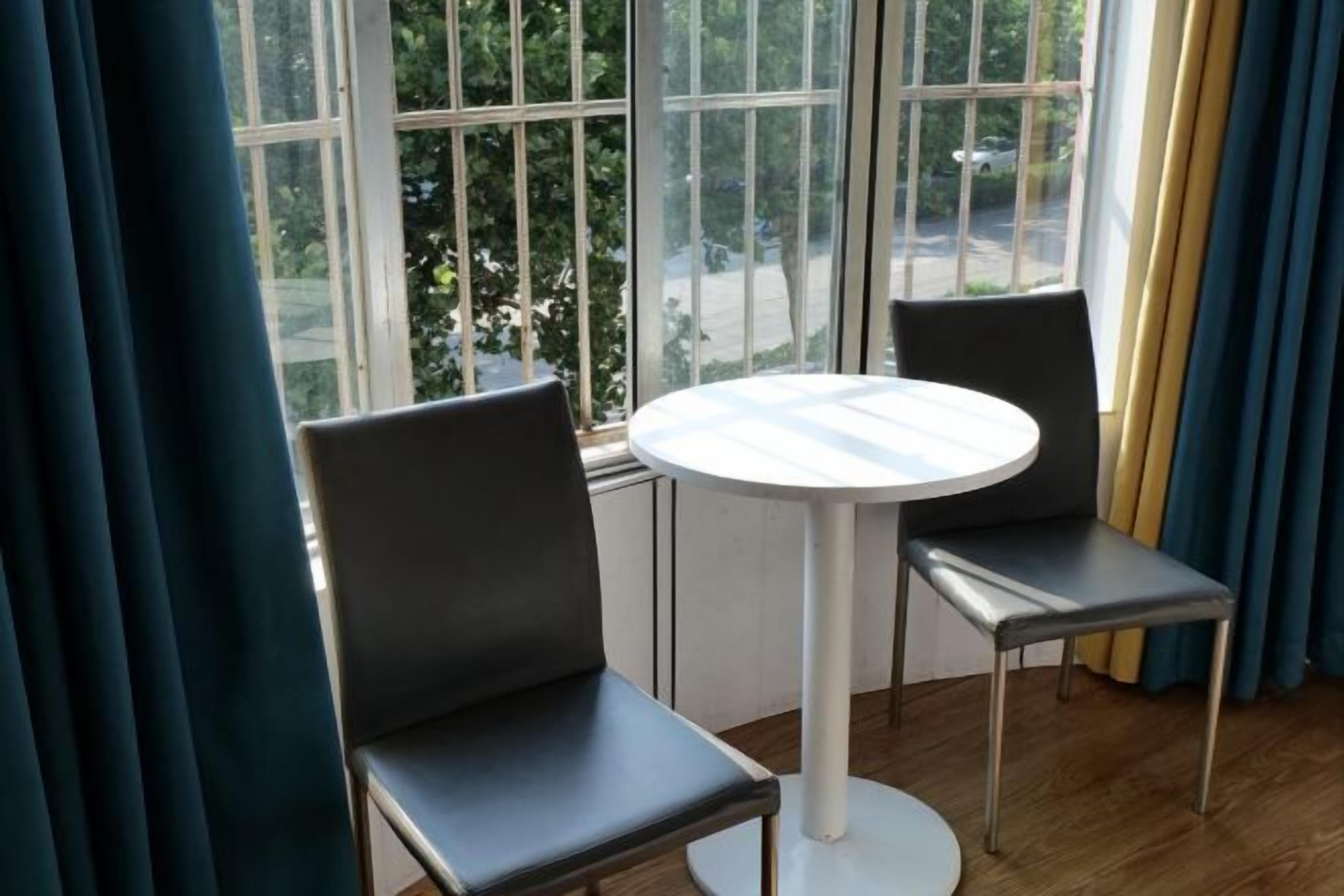 Een kamer met een ronde tafel en twee stoelen, een raam met gordijnen en uitzicht op de straat.