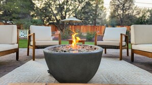 A stone fire pit with a fire burning in the center, surrounded by two white cushioned chairs and a small table.