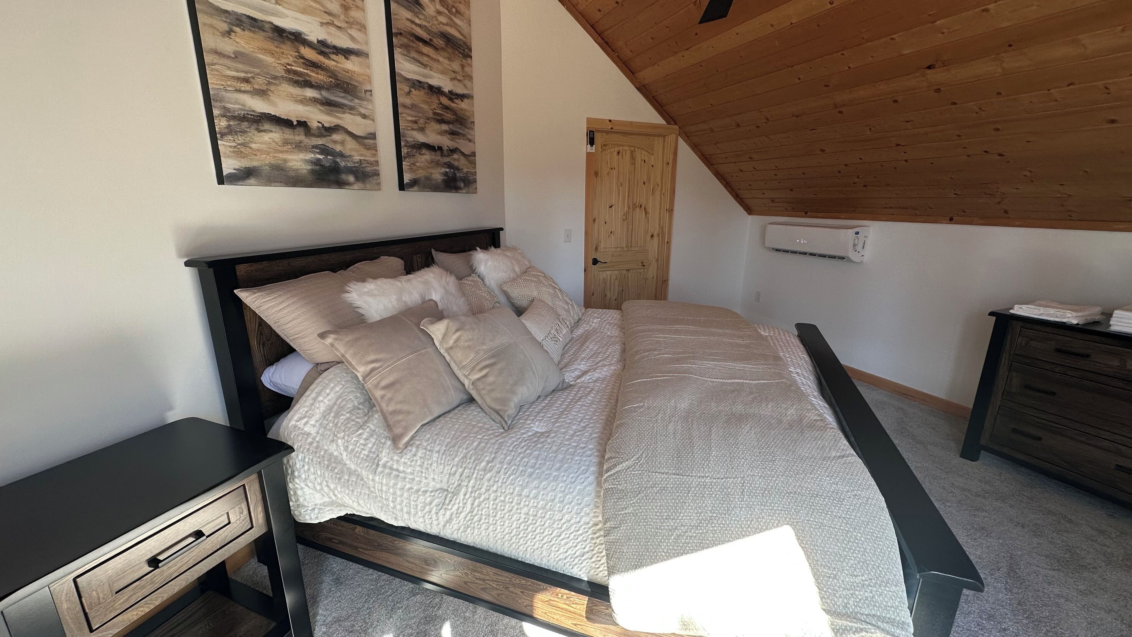 Quarto com teto de madeira, cama com várias almofadas, mesa de cabeceira, cômoda e duas pinturas na parede.
