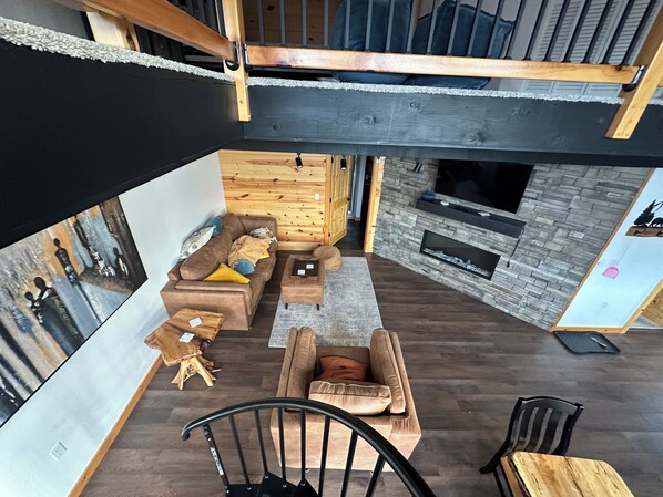 Ein modernes Wohnzimmer mit einer Couch, einem Sessel und einem kleinen Tisch. Ein Kamin und eine Holzbalkendecke sind ebenfalls vorhanden.