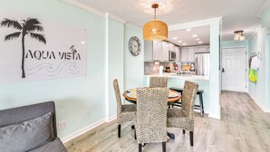 A dining area with a round table and patterned chairs, a wall clock, and a kitchen in the background.