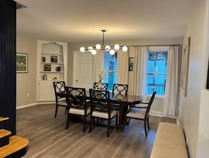 A dining area with a wooden table and chairs, a chandelier, and a view of the outside through a window with curtains. - Spacious 3bdr 2ba - walk to Breweries & Downtown (Burlington)