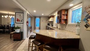 A modern kitchen with a granite countertop, wooden stools, and a dining area with a table and chairs. - Spacious 3bdr 2ba - walk to Breweries & Downtown (Burlington)