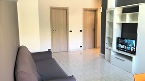 Ein modernes Wohnzimmer mit einem grauen Sofa, einem Fernseher auf einem weißen Schrank und einer Tür im Hintergrund.