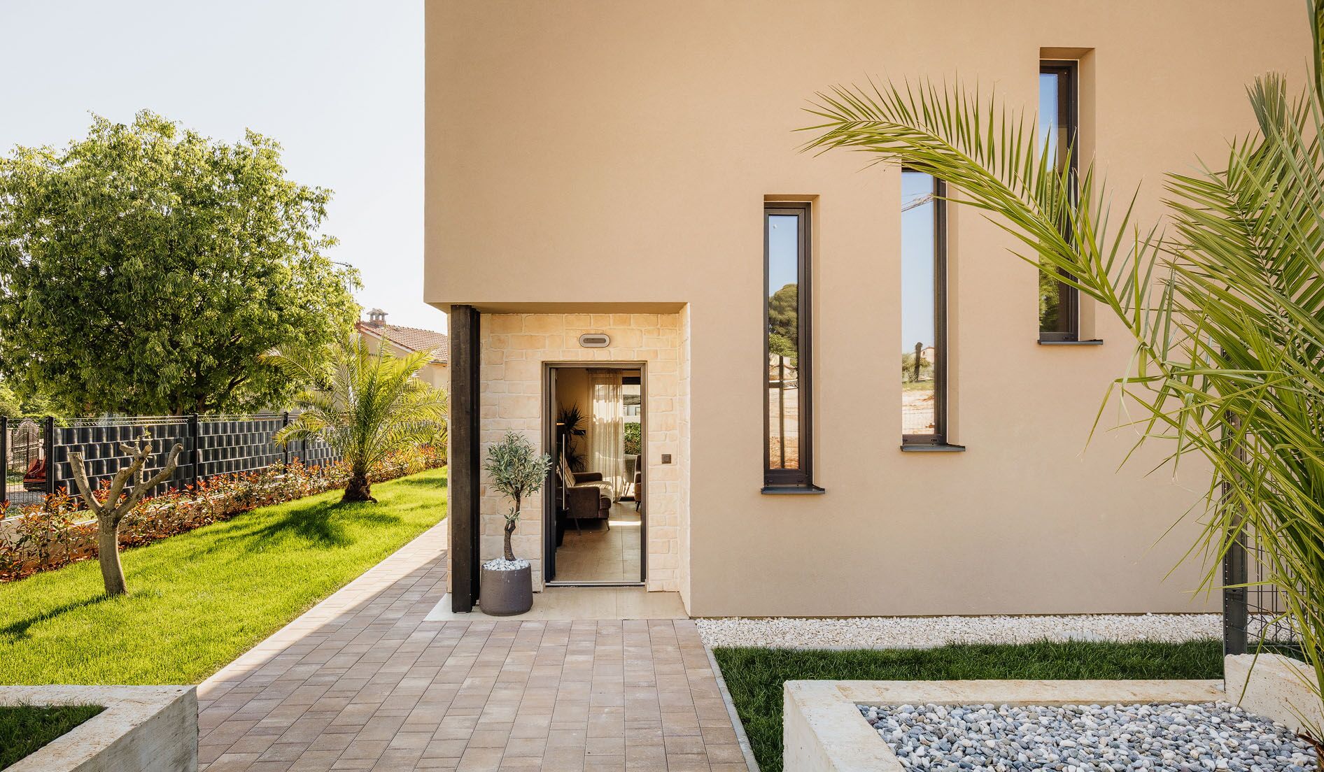 A modern building with a paved entrance, a small garden with a palm tree, and a view of the interior through a doorway.