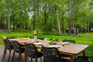 A wooden deck with a table set for a meal, surrounded by chairs, in a lush green forest setting.