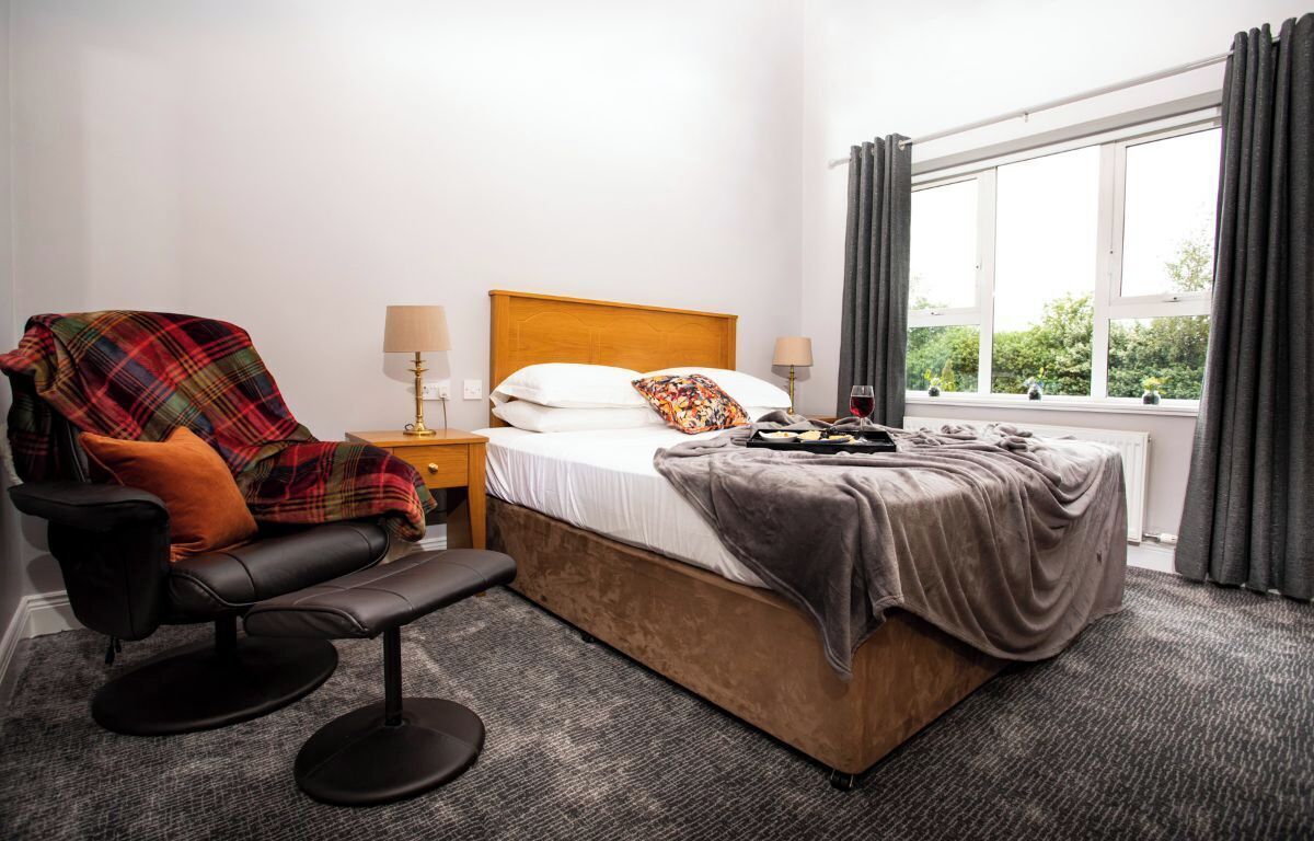 A hotel room with a bed, a chair, a bench, two bedside tables, and a window with curtains.