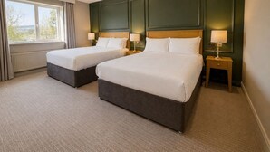 Een hotelkamer met twee bedden, een raam met gordijnen, een radiator en een nachtkastje met een lamp.