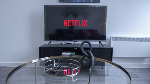 A modern living room with a flat-screen TV displaying "NETFLIX" in red letters, a black entertainment unit, and a glass coffee table with a remote control.