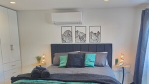 A modern bedroom with a large bed, a headboard, a small bedside table, a wall-mounted air conditioner, and a window with curtains. - Beautiful Studio w/Parking | Near CBD & Port Hills (Christchurch)