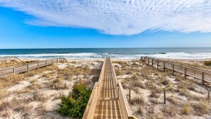 Ein hölzerner Steg führt zu einem Strand mit blauem Himmel und Blick auf den Ozean.