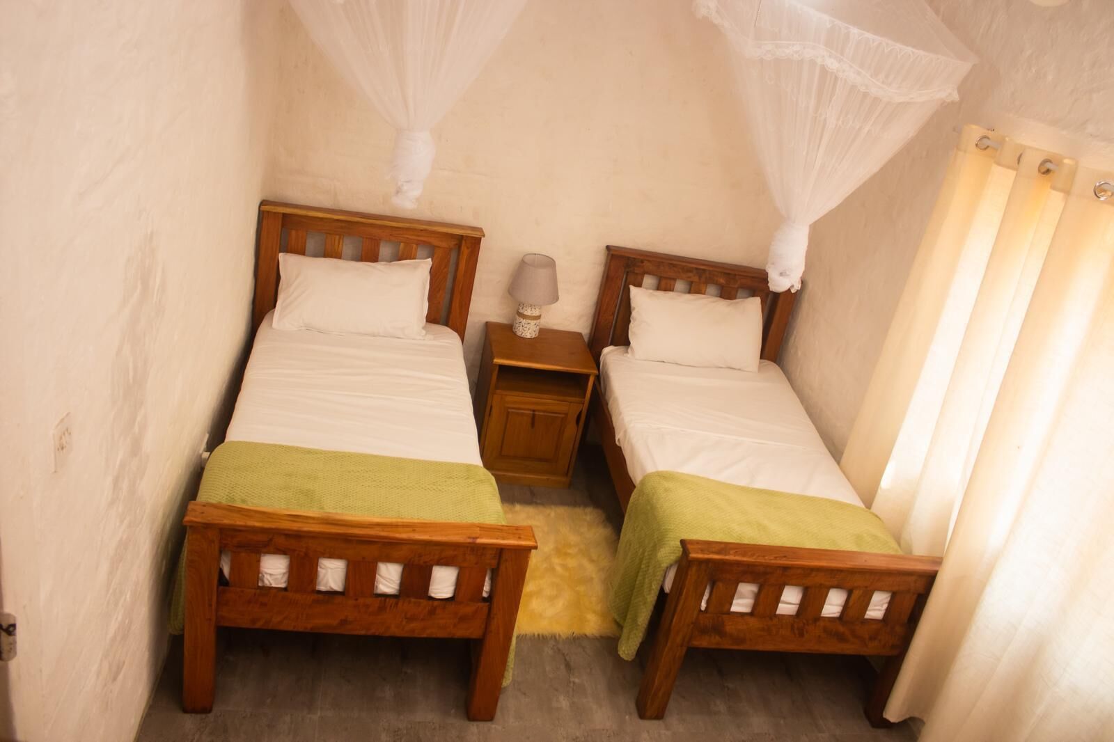Una habitación pequeña y acogedora con dos camas de madera, una mesita de noche, una lámpara y un mosquitero.