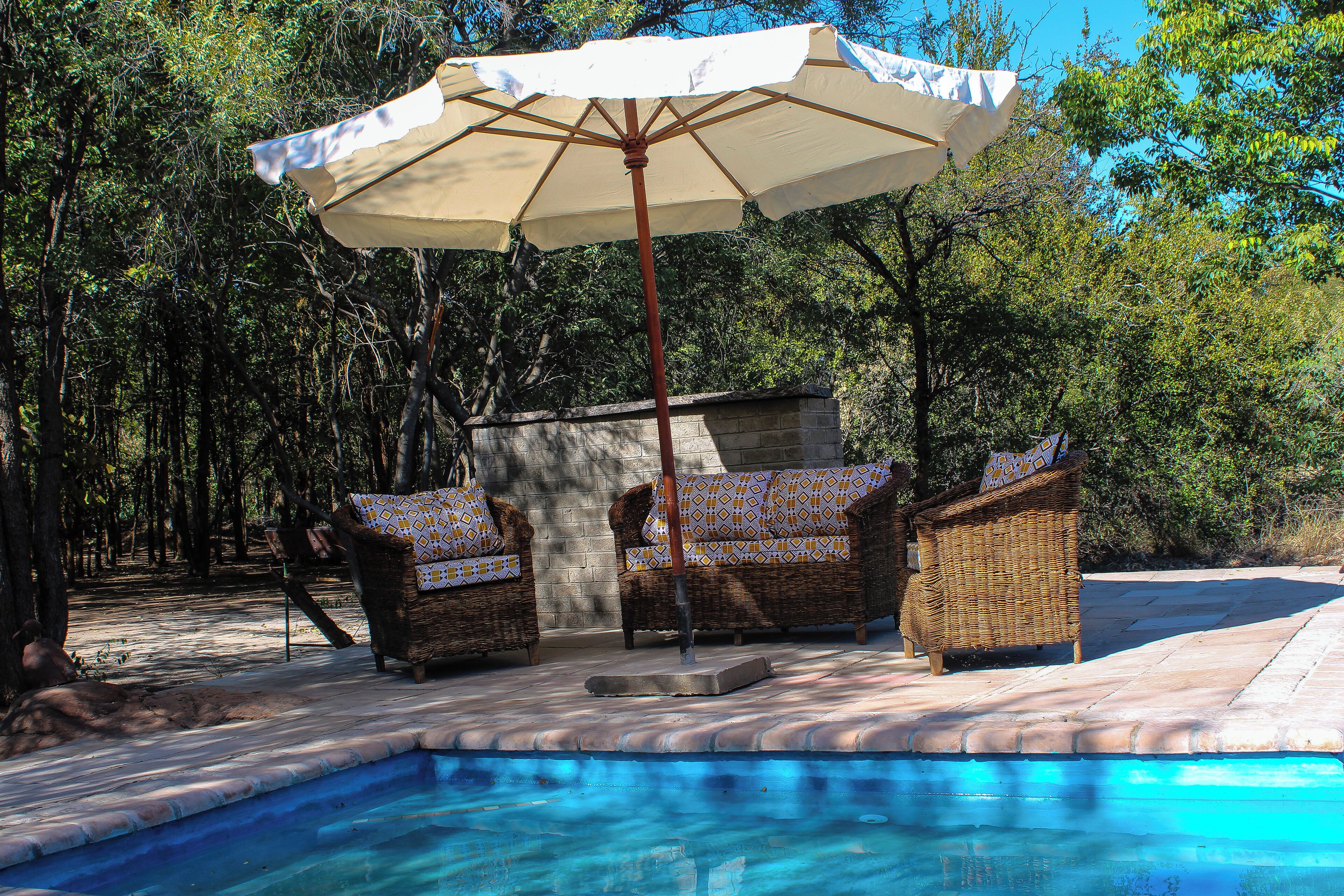 Zona de piscina con dos sillas de mimbre, un paraguas grande y un muro de piedra al fondo.