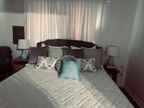 Una cama bien hecha con cubrecama acolchado, flanqueada por dos mesitas de noche con lámparas, y un cabecero con cojines estampados.