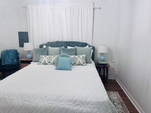 A neatly made bed with a quilted white cover, flanked by two bedside tables with lamps, a blue blanket on a chair, and a patterned rug on the floor.