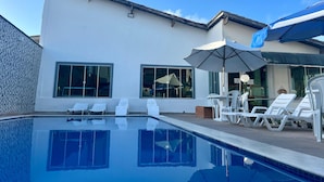 Una piscina con sillones de descanso y sombrillas, un edificio blanco con grandes ventanales y un cielo azul.