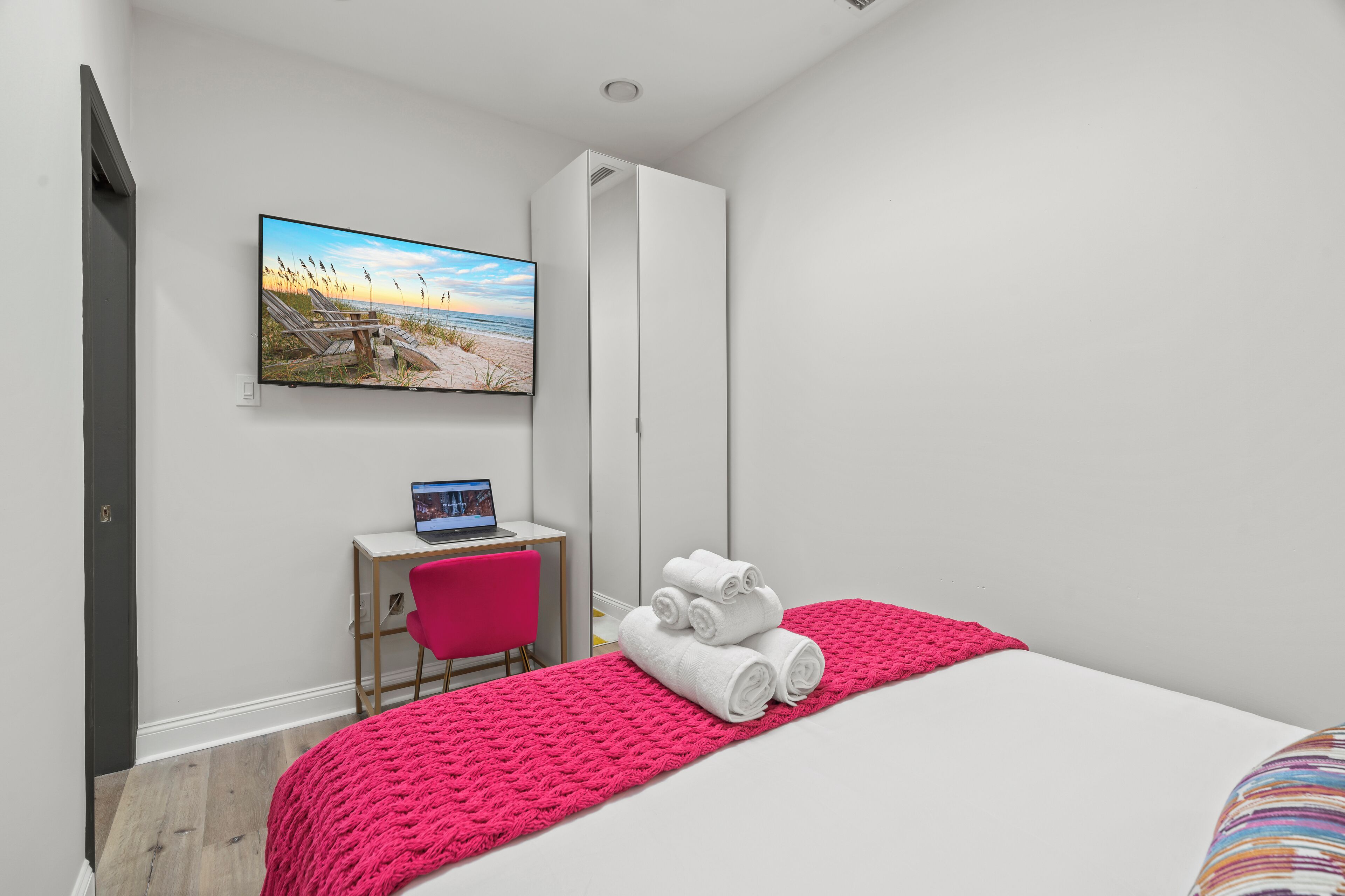 A hotel room with a bed, a desk with a laptop, a chair, a TV on the wall, and a window with a beach view.