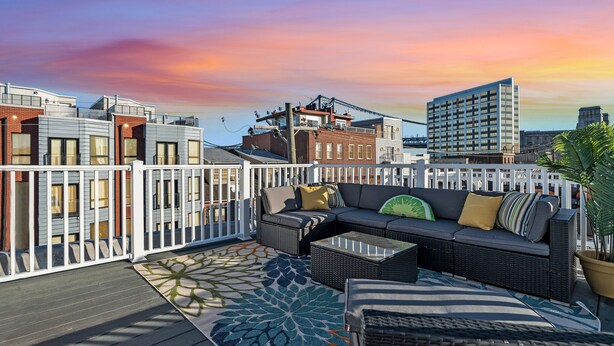 Una terraza en la azotea con valla blanca, zona de estar al aire libre y vista del horizonte urbano al atardecer.
