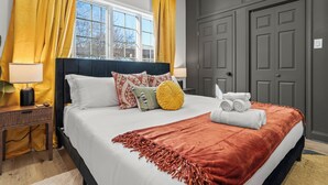 Una cama bien tendida con una colcha blanca y naranja, un cabecero oscuro y almohadas decorativas. Hay una mesita de noche con una lámpara y una pequeña mesa junto a la cama.