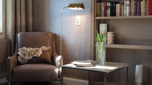 A brown leather armchair with a decorative pillow, a floor lamp, a small table with a book, a vase with green plants, and a bookshelf with books.
