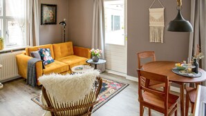 Ein gemütliches Wohnzimmer mit einem senfgelben Sofa, einem weißen, flauschigen Sessel, einem Holzespeisetisch mit Stühlen, einem kleinen Beistelltisch mit Blumen in einer Vase und einer Wandbehagung aus Makramee.
