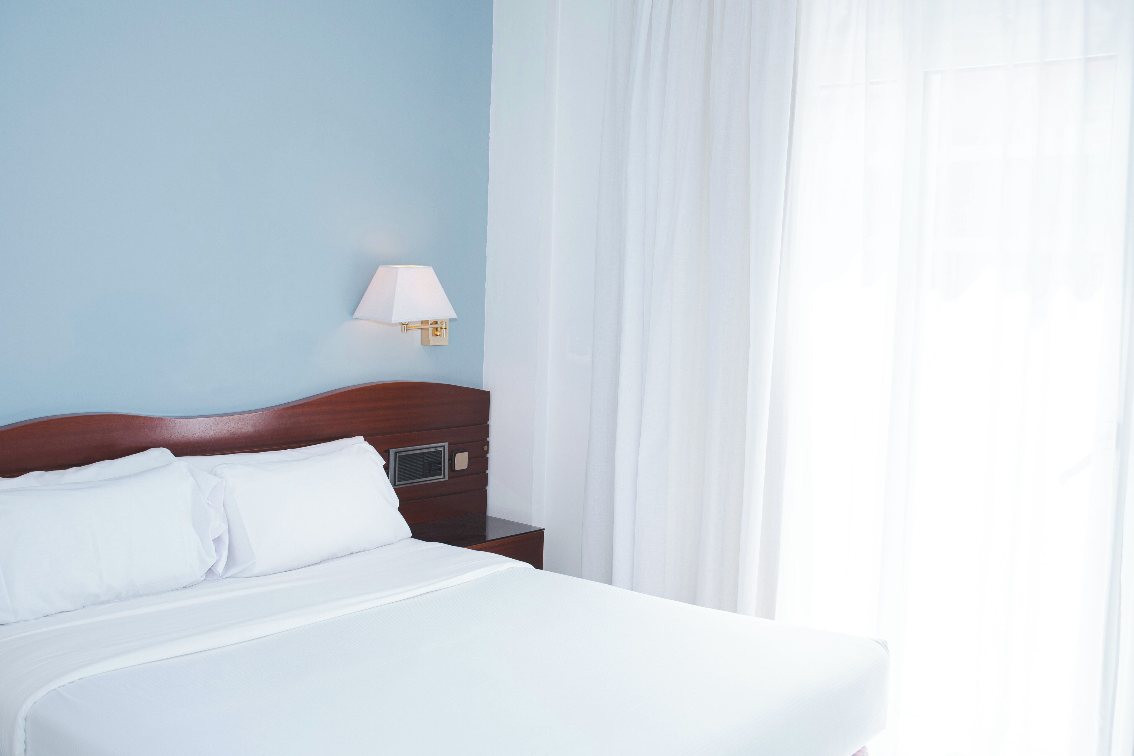 Habitación de hotel con una cama bien hecha, cabecera de madera, mesita de noche con lámpara y ventana con cortinas traslúcidas.