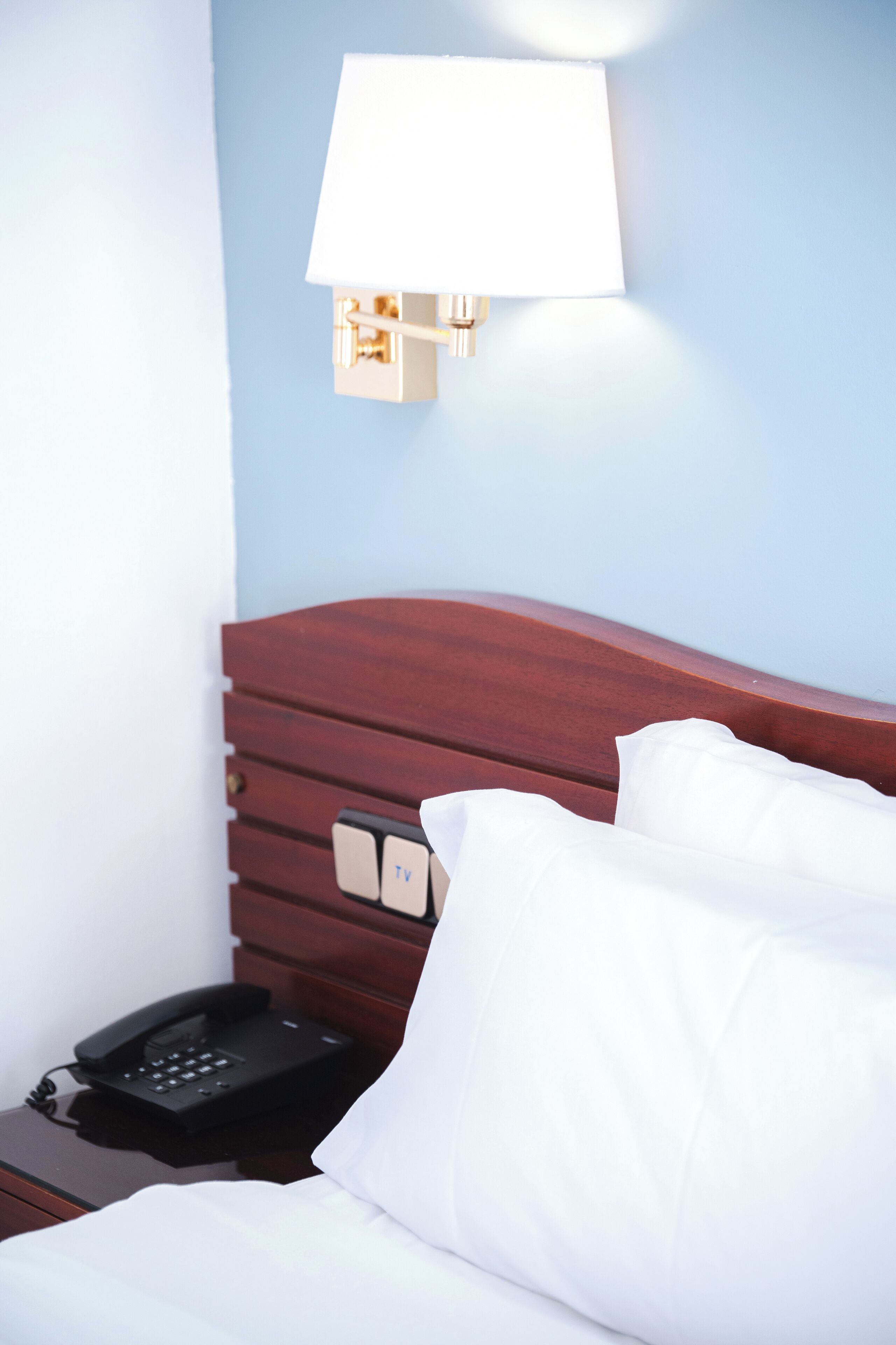 Habitación de hotel con cabecera de madera, teléfono en la mesita de noche, lámpara y ropa de cama blanca.