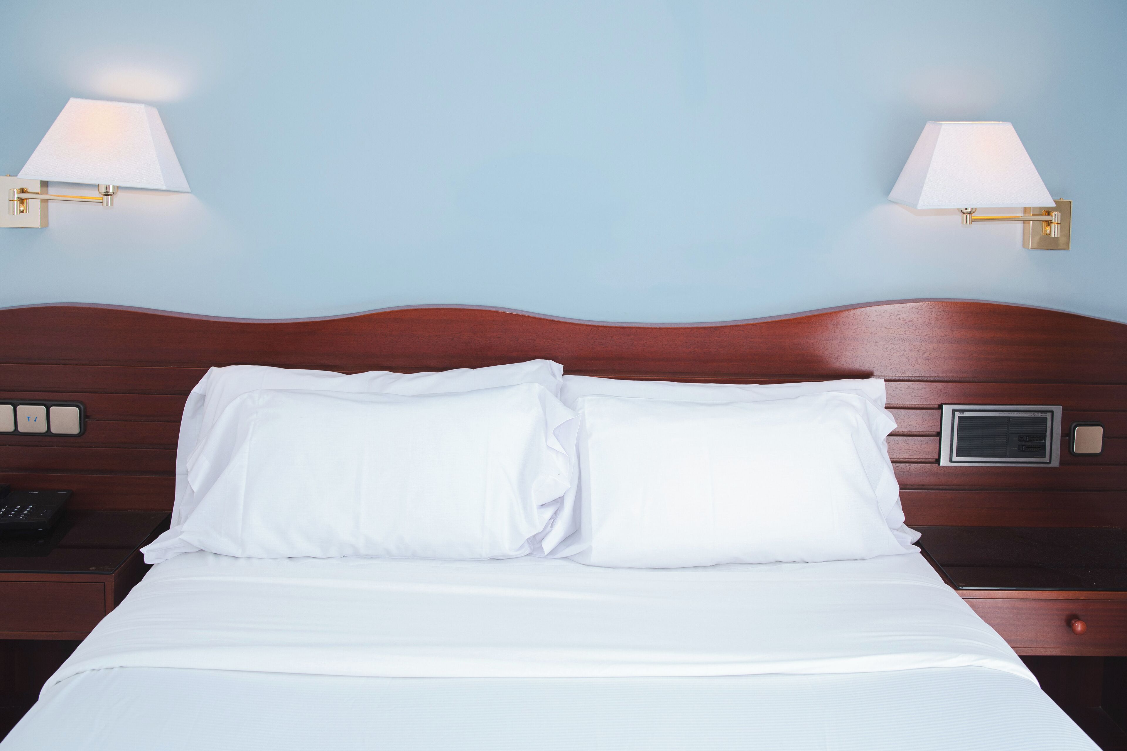 A neatly made bed with white linens, a wooden headboard, and two wall-mounted lamps.