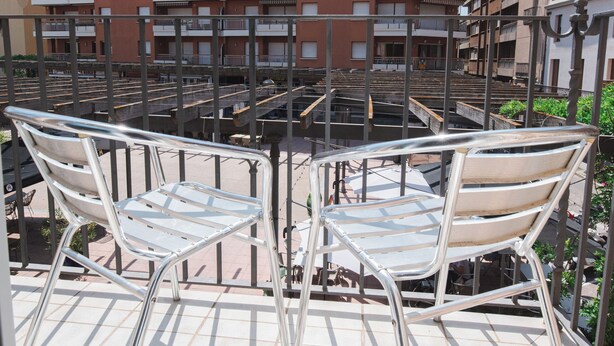 Un balcón con dos sillas blancas de metal y una mesa, con vistas a un edificio residencial de varios pisos con balcones.