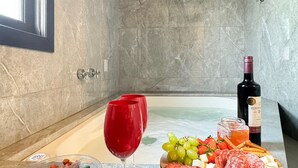 A room with a marble floor, a bathtub, a wine bottle, a glass of red wine, and a platter of food including grapes, cheese, and meats. - Casa com piscina Praia da Vila Nova (Imbituba)