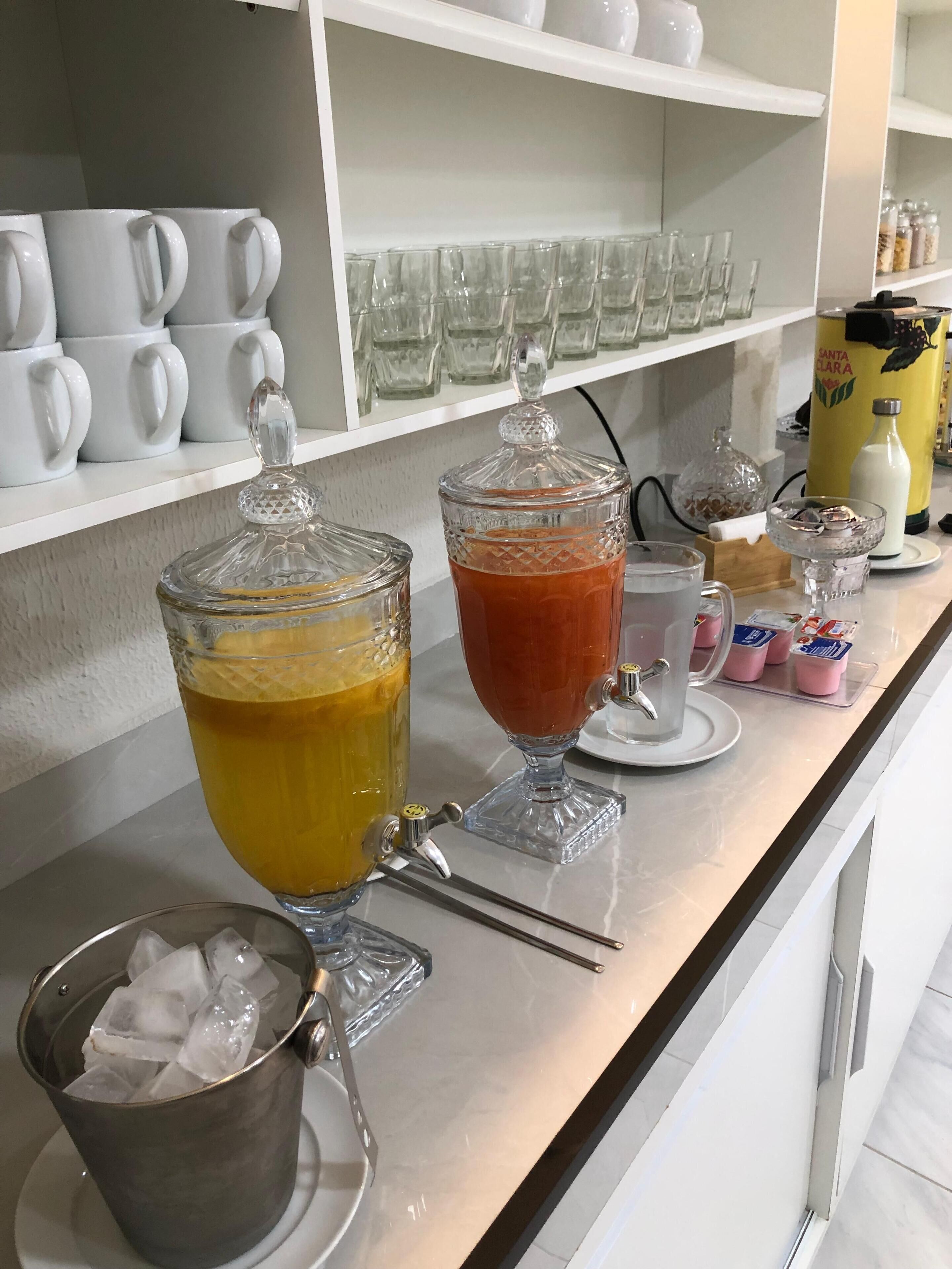 A bar area with two large dispensers of orange and red beverages, a metal bucket with ice, and a variety of glassware on shelves.