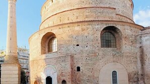 Un edificio histórico con una torre redonda y ventanas de arco, rodeado de áreas verdes y ruinas.