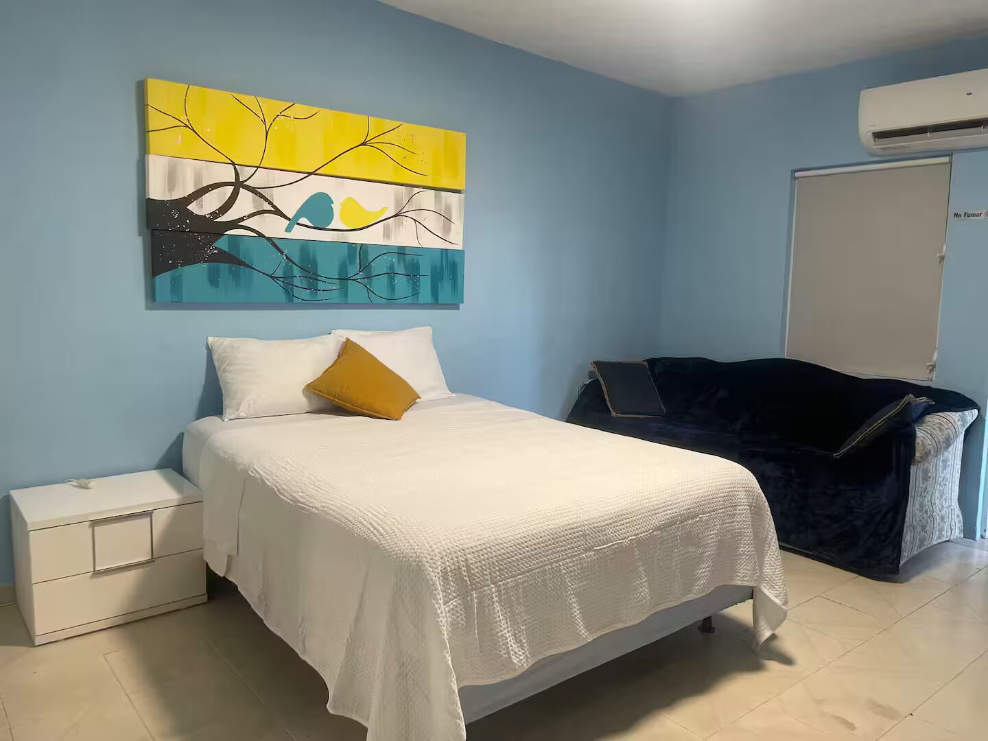 A bedroom with a bed, a nightstand, a dresser, a blue couch, and a wall painting.