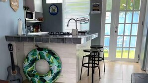 A kitchen with a built-in island, a dining area with a bar stool, a guitar, and a green leaf-patterned cushion. - The Retreat - 5 min to Airport (Carolina)