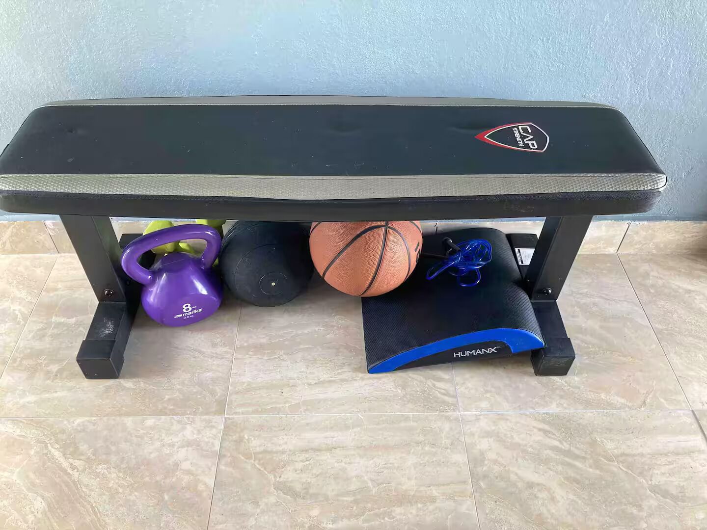 Un banc de musculation orné d’un logo, un haltère russe violet, un ballon lesté noir, un ballon de basket et un élastique de résistance bleu.