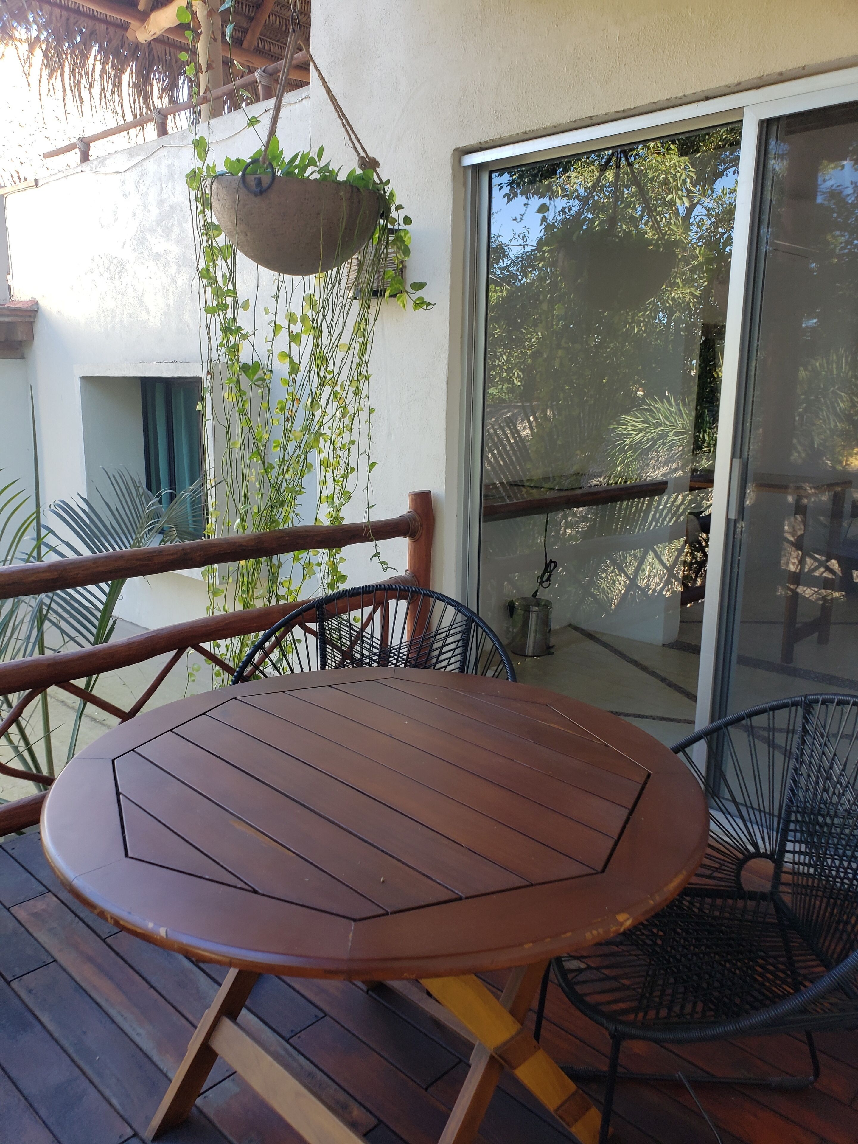 A wooden table and chairs on a balcony with a hanging plant, a sliding glass door, and a view of trees.