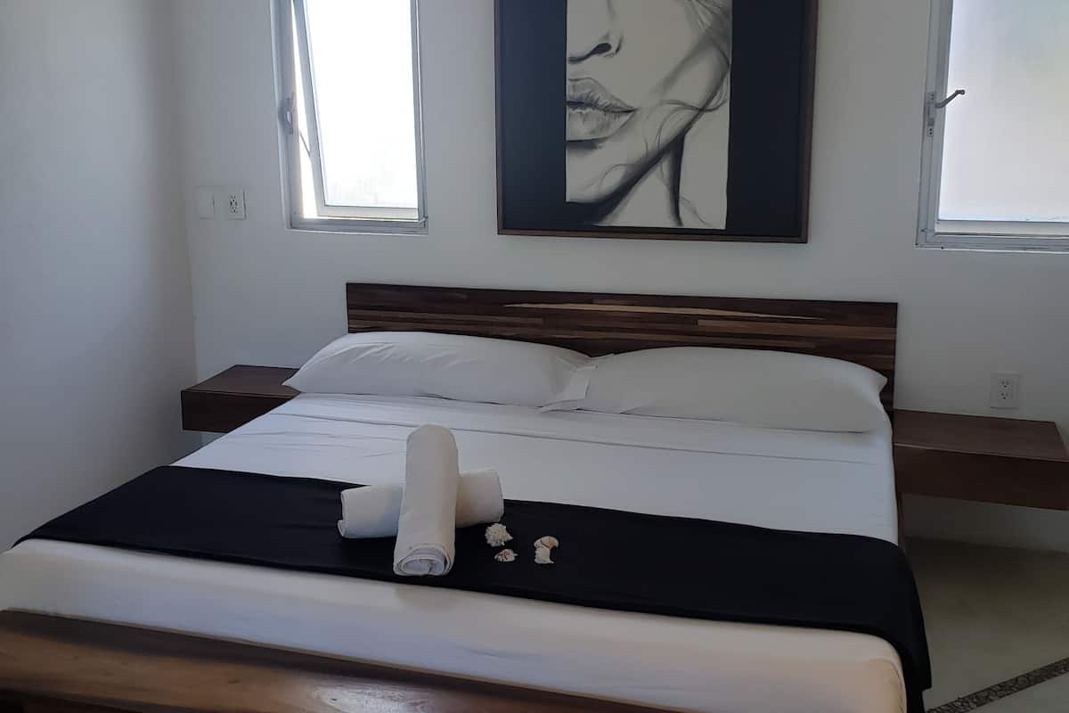 A bedroom with a large bed, two bedside tables, a wall-mounted artwork, and two windows.