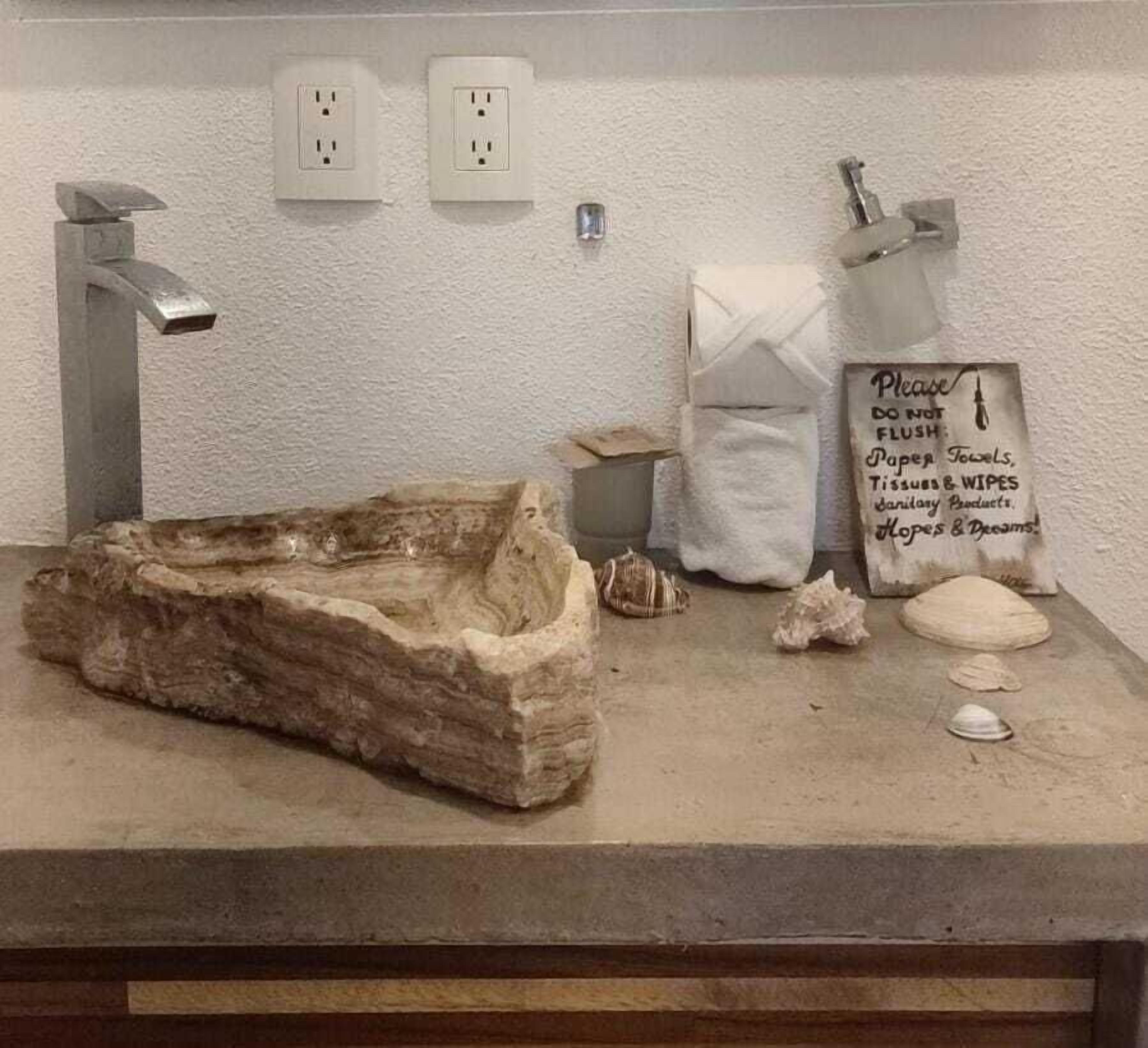 A bathroom with a stone basin, two electrical outlets, a towel dispenser, and a sign instructing not to flush paper towels, tissues, and wipes.