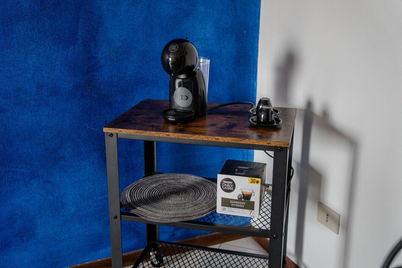 A small wooden table with a coffee maker, a cup, and a box of Nestle Dolce Gusto espresso.