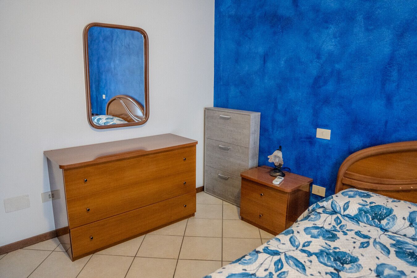 Una camera da letto con una cassettiera in legno, un piccolo mobiletto, un comodino con una lampada e un letto con copriletto floreale.